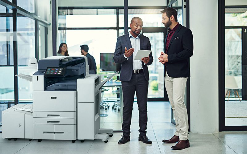 Deux hommes dans un bureau discutent sur une tablette à côté d'une imprimante multifonction couleur Xerox® AltaLink® série C8200